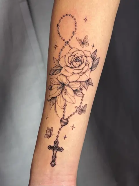 A rosary of rose, lily, butterflies, and a cross, falling down the forearm.